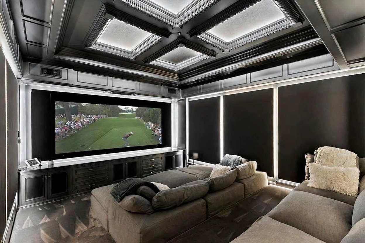 This is a luxurious home theater room featuring a large screen displaying a golf game, a modular gray sectional sofa, and a sophisticated black coffered ceiling with recessed lighting. The room is designed for immersive entertainment, with dark walls and controlled lighting to enhance the viewing experience. The overall impression is one of comfort and high-end design.