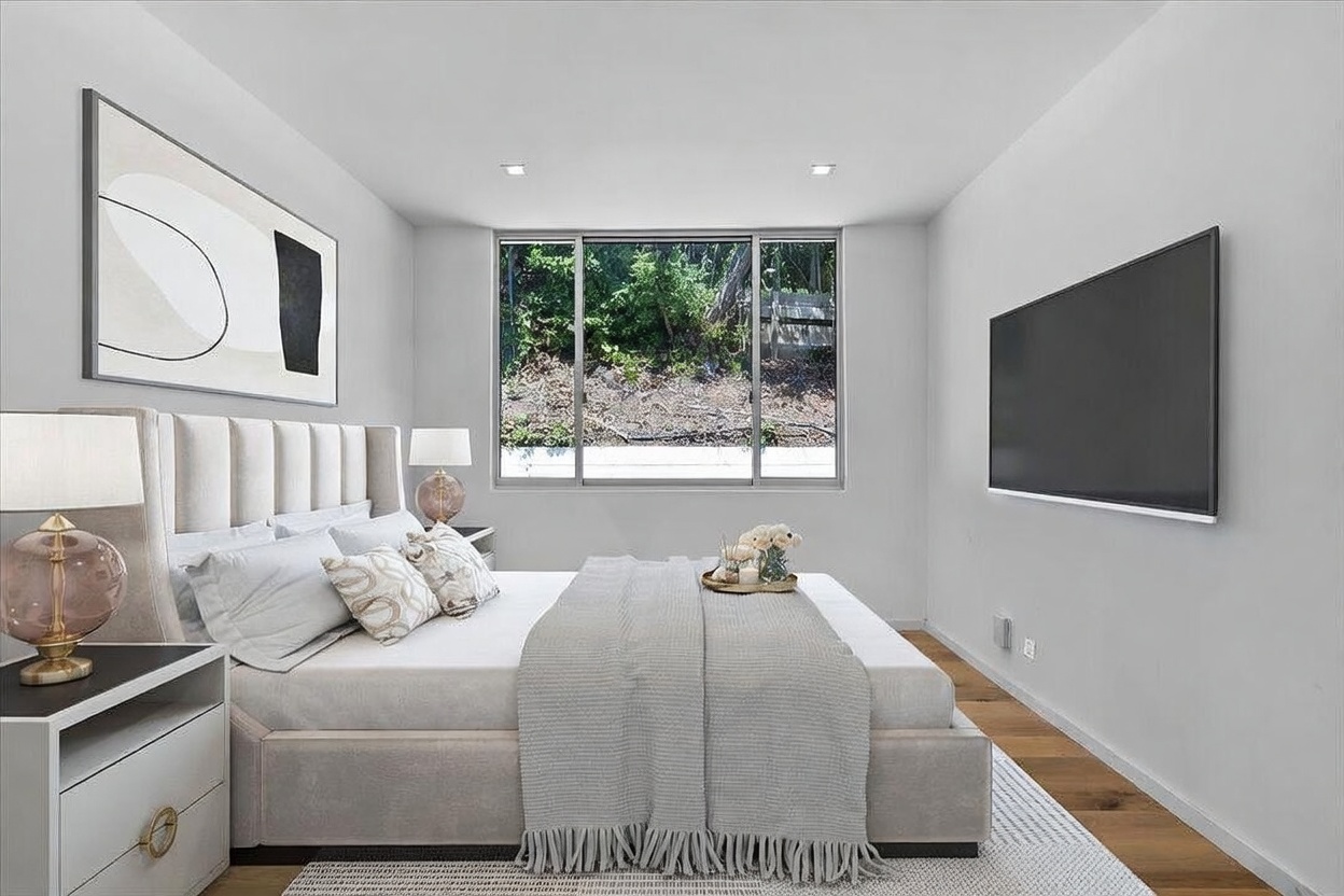 This modern primary bedroom features a neutral color palette with a plush, upholstered headboard and a neatly made bed layered with textured linens. A large window centered on the back wall provides a view of the natural landscape outside, while a wall-mounted television is positioned opposite the bed. The room is accented by contemporary bedside lamps and a large piece of abstract wall art, creating a serene and sophisticated atmosphere.