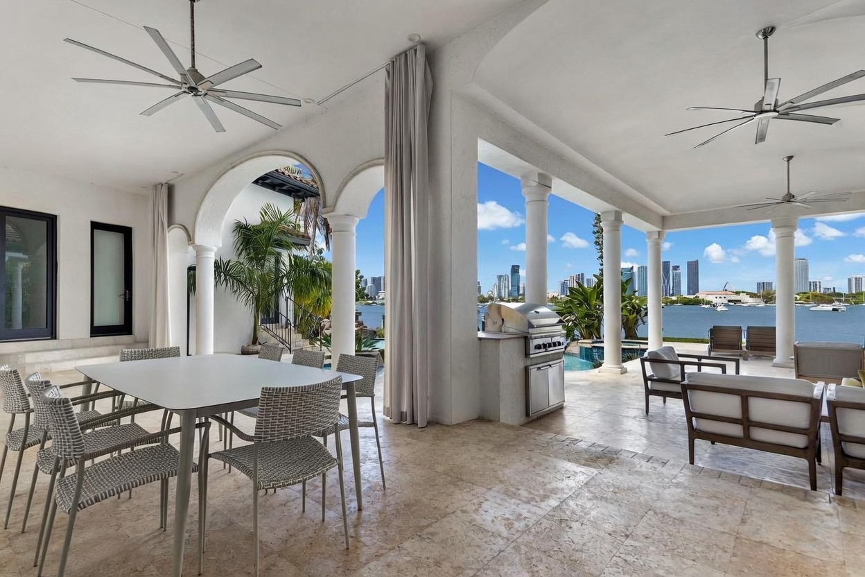 This expansive covered patio offers a luxurious outdoor living space with stone flooring and multiple ceiling fans. The area features a dining set, a built-in outdoor grill station, and comfortable lounge seating, all overlooking a waterfront view with a city skyline in the distance. The open-air design, framed by elegant columns and arches, creates a seamless transition between the indoor and outdoor environments.
