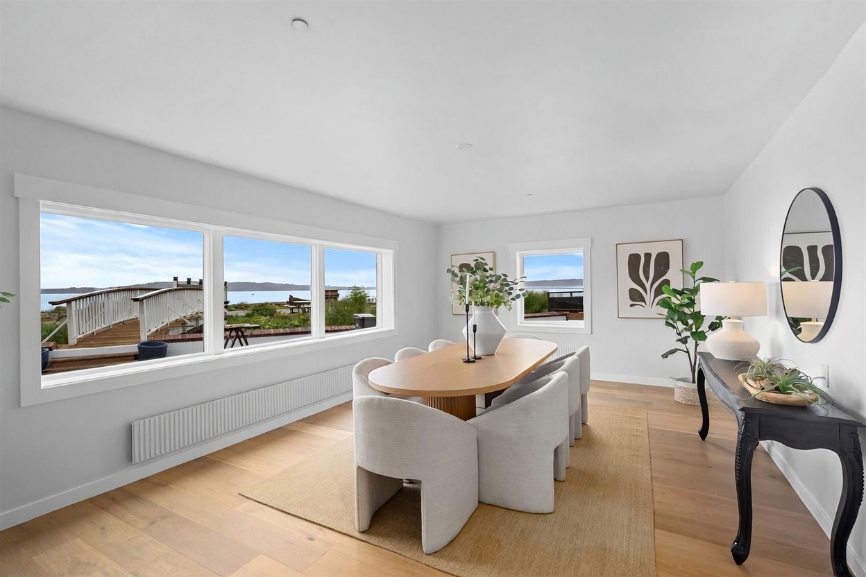 This bright and airy dining room features a modern oval wooden table surrounded by six upholstered chairs, set atop a textured area rug. Large windows along the wall provide expansive views of the water and an outdoor deck, filling the space with natural light. A sleek black console table with a round mirror and a potted plant adds a sophisticated touch to the minimalist, light-toned interior.