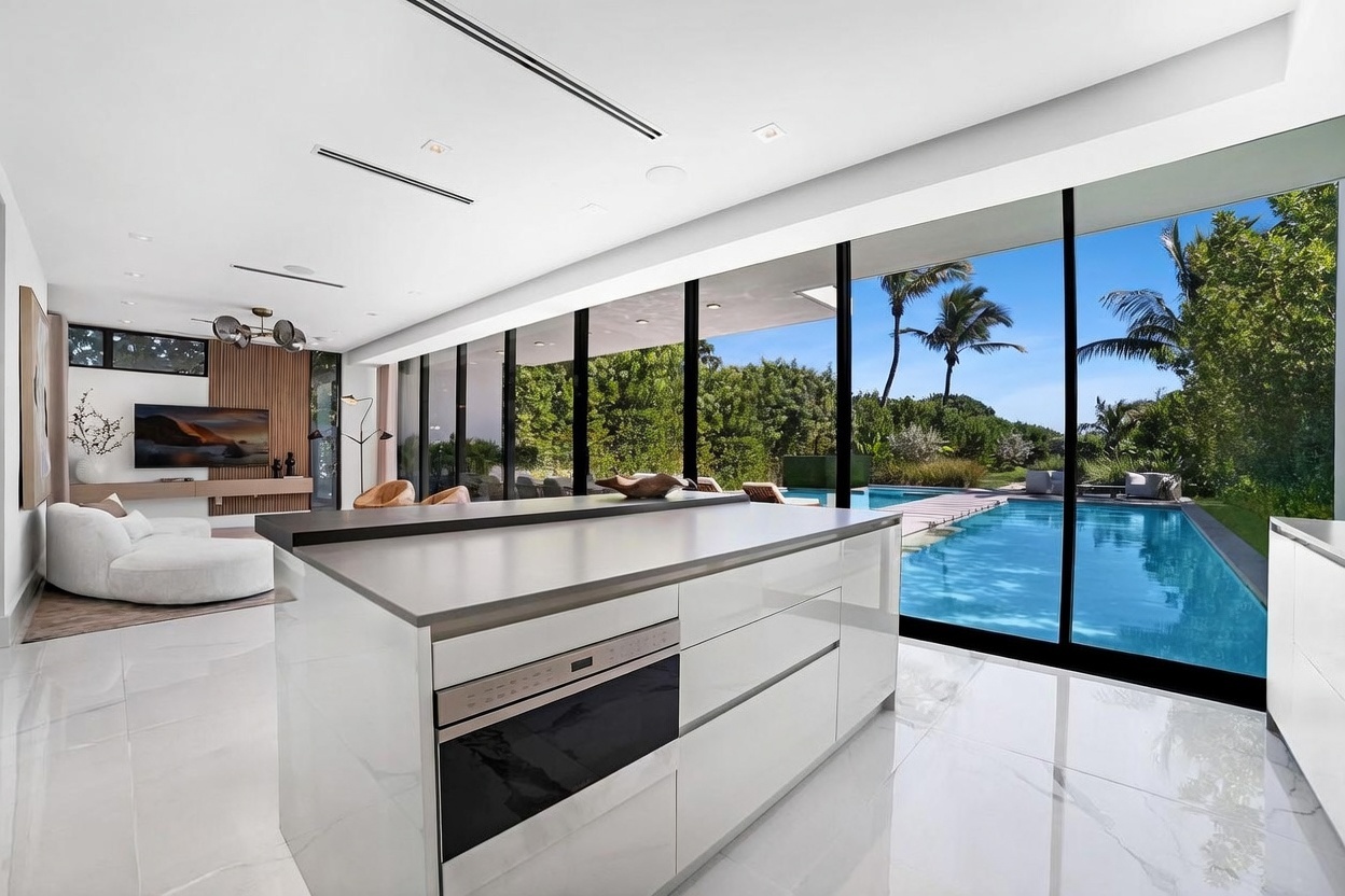 This modern, open-concept kitchen features a sleek white island with a built-in oven and a dark countertop, seamlessly transitioning into a bright living area. Large, floor-to-ceiling windows provide an expansive view of the outdoor swimming pool and lush greenery, creating a strong indoor-outdoor connection. The space is characterized by clean lines, glossy surfaces, and a minimalist aesthetic that emphasizes natural light and luxury.