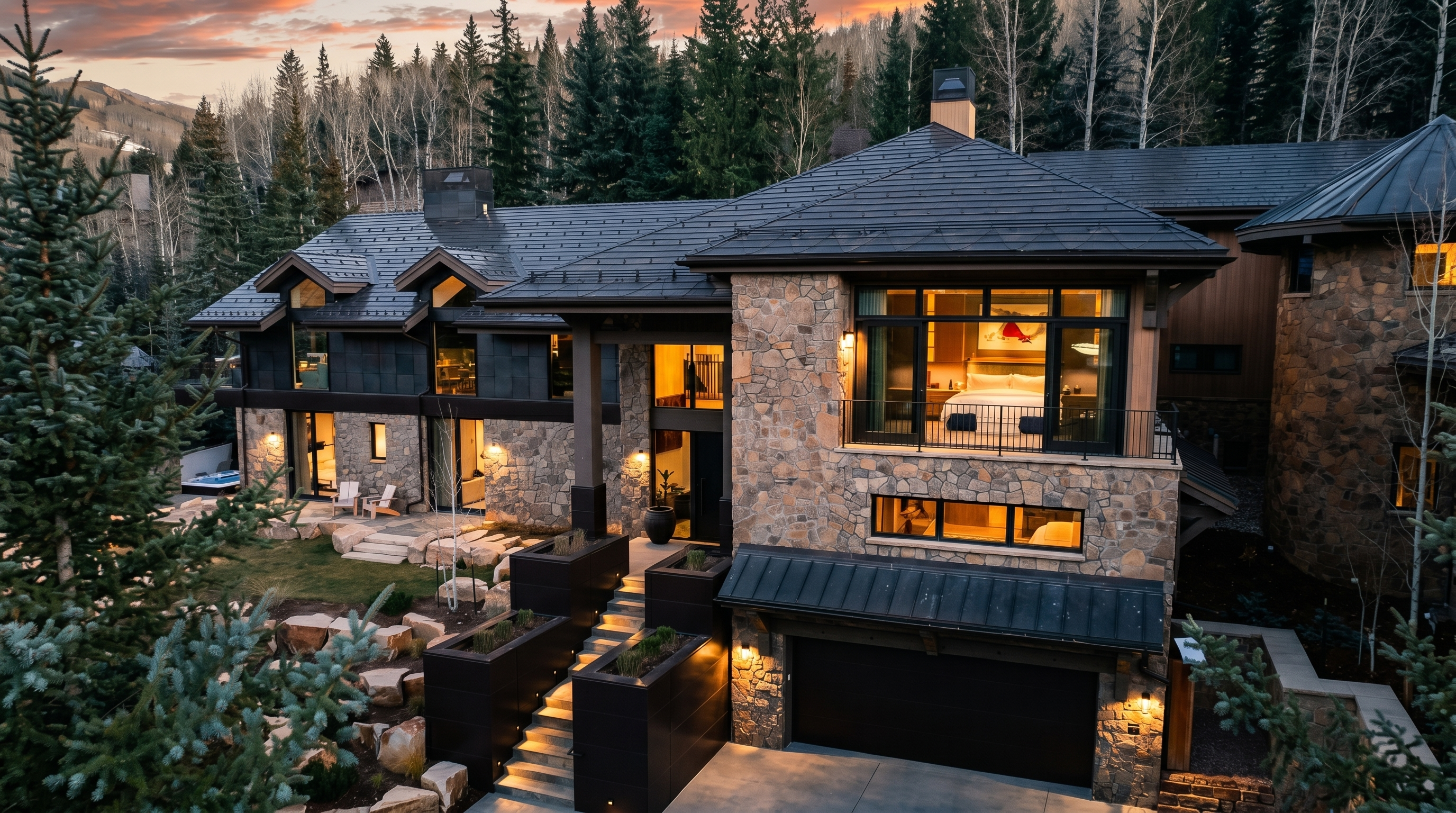 This striking mountain residence features a sophisticated blend of natural stone and dark metal siding, set against a backdrop of evergreen trees and a vibrant sunset. The multi-level design includes a prominent garage, a stone-paved driveway, and a tiered entryway with modern planters leading to the main door. Large windows, including a balcony off an upper-level bedroom, provide a seamless connection to the surrounding alpine landscape.