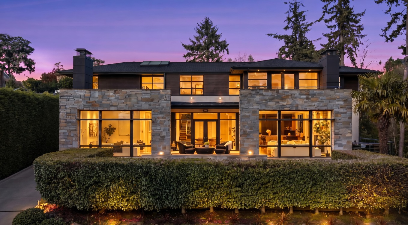 This contemporary two-story residence features a striking combination of dark horizontal siding and textured stone cladding. The rear facade is defined by expansive glass walls that provide a seamless connection to the interior living spaces, while a manicured hedge and tiered landscaping frame the foreground. The twilight setting highlights the warm interior lighting, creating an inviting and sophisticated atmosphere.