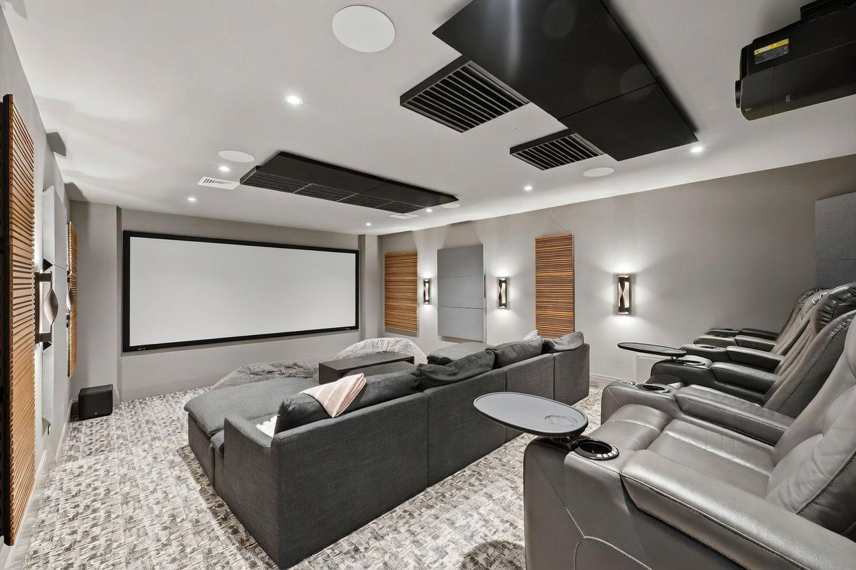 This dedicated home theater room features a large projection screen, tiered seating with plush gray recliners, and a comfortable sectional sofa. The space is acoustically treated with wall-mounted panels and ceiling-mounted sound baffles, creating an immersive cinematic environment. The room is finished with neutral gray walls, patterned carpeting, and modern wall sconces that provide soft, ambient lighting.
