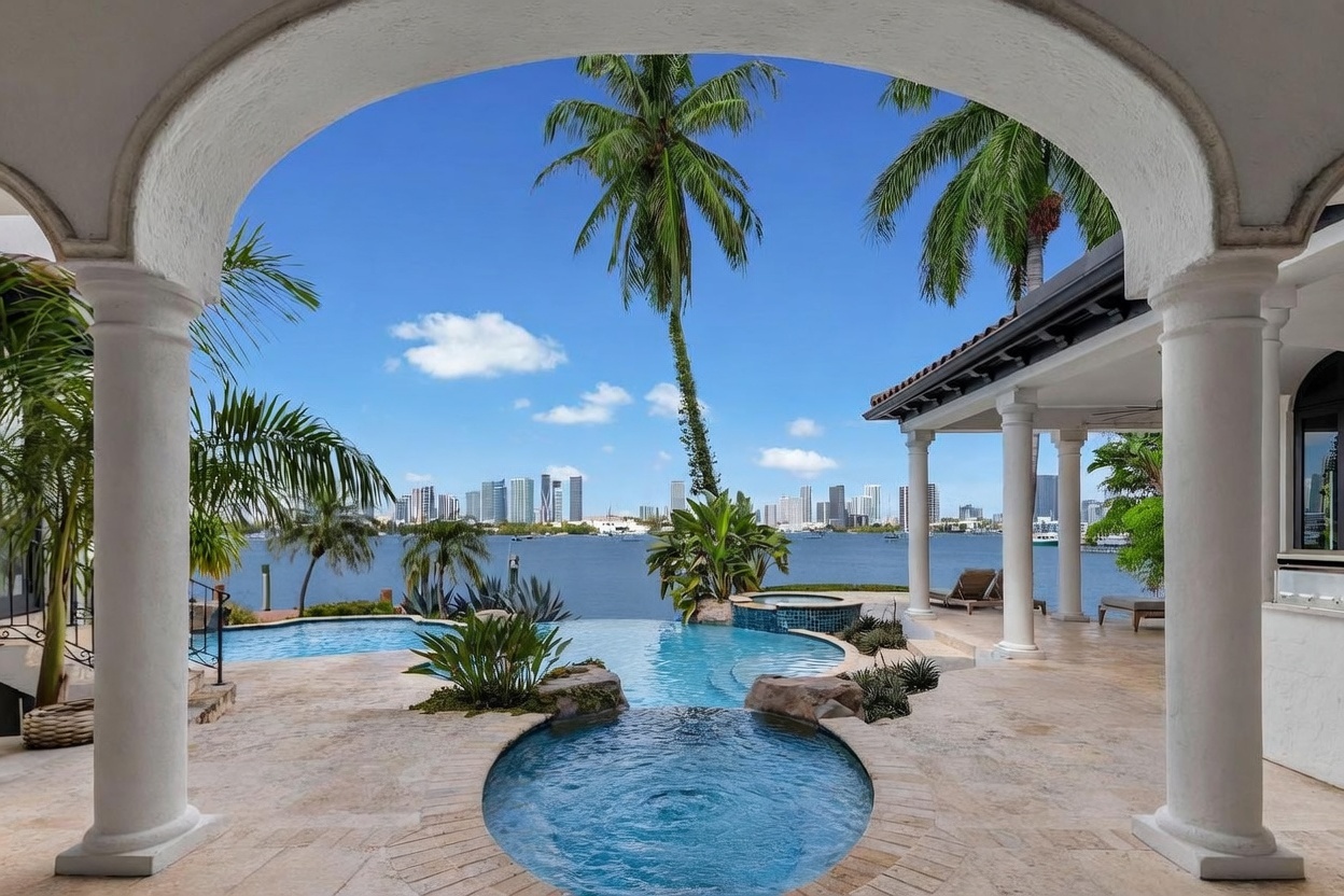 This luxurious outdoor living space features a multi-tiered infinity-edge pool and spa that seamlessly blend into the expansive waterfront view. Framed by a classic arched stone colonnade, the patio offers a serene vantage point overlooking a calm bay with a distant city skyline. The scene is bathed in bright, natural sunlight, highlighting the elegant stone decking and lush tropical landscaping.