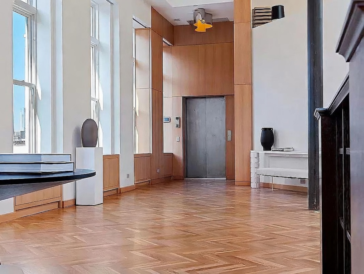 This interior view captures a spacious, high-ceilinged hallway featuring rich, herringbone-patterned wood flooring and light-toned wood paneling. A central elevator with metallic doors serves as a focal point, flanked by minimalist decor including a pedestal-mounted vase and a sleek console table. The perspective is centered, emphasizing the architectural symmetry and the clean, sophisticated aesthetic of the space.