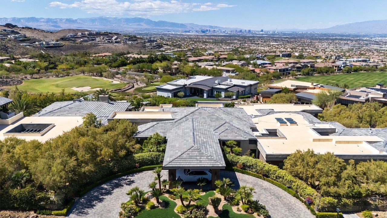 This high-angle aerial perspective captures a sprawling, modern luxury estate nestled within a desert landscape. The home features a complex, multi-gabled roofline with grey tiles, a circular paved driveway leading to a covered porte-cochère, and lush, manicured landscaping. In the background, a vast residential community stretches toward a distant mountain range under a clear blue sky.