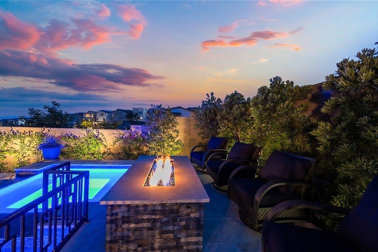 This inviting outdoor living space features a stone-clad fire pit as the centerpiece, surrounded by comfortable dark-cushioned armchairs. To the left, a swimming pool with a safety fence is visible, while lush greenery and a stone wall provide privacy. The scene is captured at dusk, with a vibrant sunset sky casting a warm glow over the patio area.