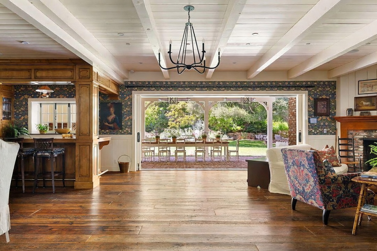 This spacious living area features warm wood flooring and a white beamed ceiling, creating a rustic yet refined atmosphere. Large, open glass doors provide a seamless transition to an outdoor dining patio, while a patterned armchair and a bar area with stools add character to the room. The space is accented by floral wallpaper and a classic chandelier, offering a welcoming and airy feel.