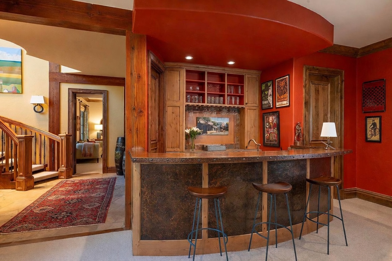 This basement area features a custom built-in wet bar with a copper-toned countertop and a dark, textured front panel, complemented by three bar stools. The space is defined by bold red walls, a curved soffit above the bar, and warm wood trim, creating a cozy and inviting atmosphere. A staircase with a wooden railing leads to an adjacent room, while framed artwork adds a personalized touch to the vibrant living space.