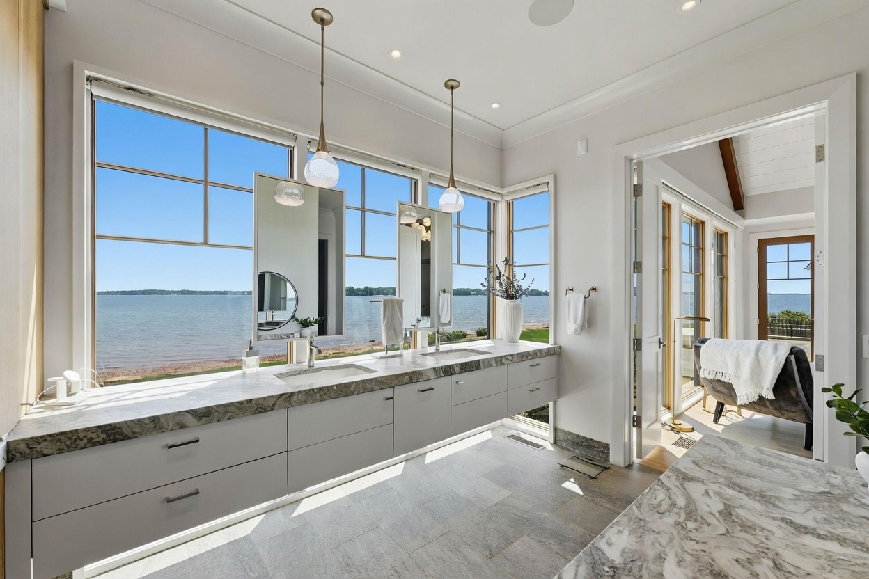 This primary bathroom features a long, floating vanity with a stone countertop and dual sinks, positioned directly in front of large windows that offer a panoramic view of the lake. The space is bright and airy, with light-toned cabinetry, neutral tile flooring, and elegant pendant lighting hanging above the vanity. A doorway leads to an adjacent room, creating a seamless flow between the bathroom and the rest of the home.