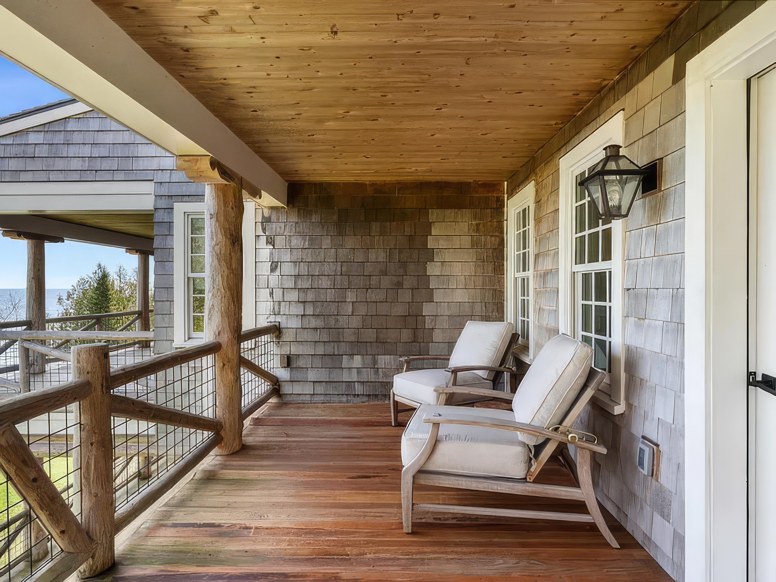 This covered balcony features rustic wood-plank flooring and a natural wood ceiling, creating a warm, inviting outdoor living space. Two comfortable lounge chairs with light-colored cushions are positioned to overlook the water, while a sturdy log-post railing with wire mesh provides safety without obstructing the view. The exterior walls are finished in weathered wood shingles, complemented by a classic wall-mounted lantern light fixture.
