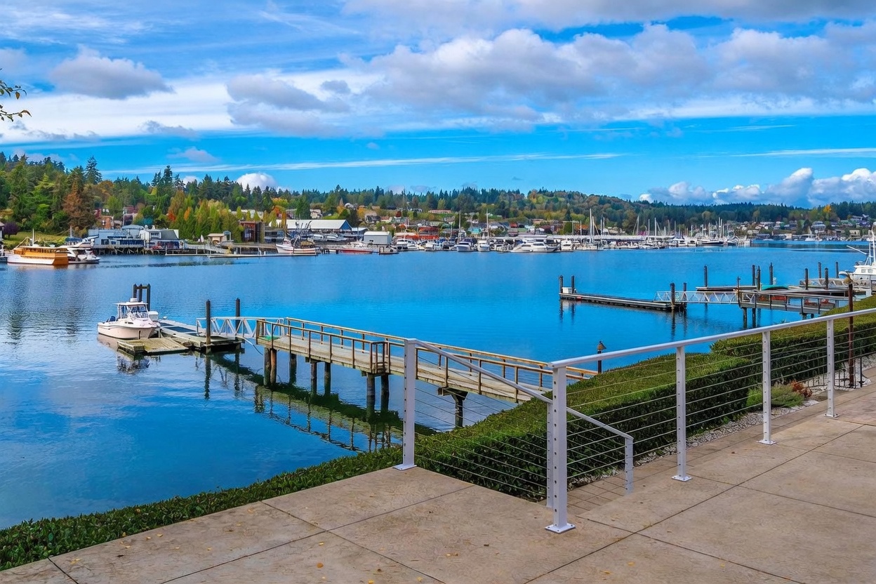 This image captures a scenic waterfront view from a paved patio area, featuring a modern metal railing with cable infill. A wooden pier extends into the calm blue water, where a small boat is docked, while a bustling marina filled with numerous boats and a backdrop of lush, tree-covered hills completes the picturesque landscape. The perspective is from an elevated vantage point, emphasizing the serene connection between the property and the harbor.