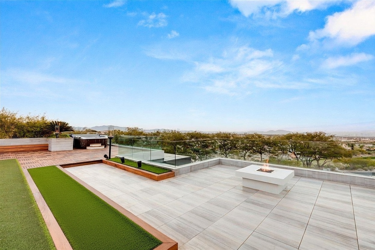 This expansive rooftop terrace offers a sophisticated outdoor living space with panoramic views of the surrounding landscape. The area features light-toned tile flooring, a modern square fire pit, and a built-in hot tub set against a backdrop of lush trees and distant mountains. A glass railing provides an unobstructed view, while a landscaped planter bed with red flowers adds a touch of natural color to the contemporary design.
