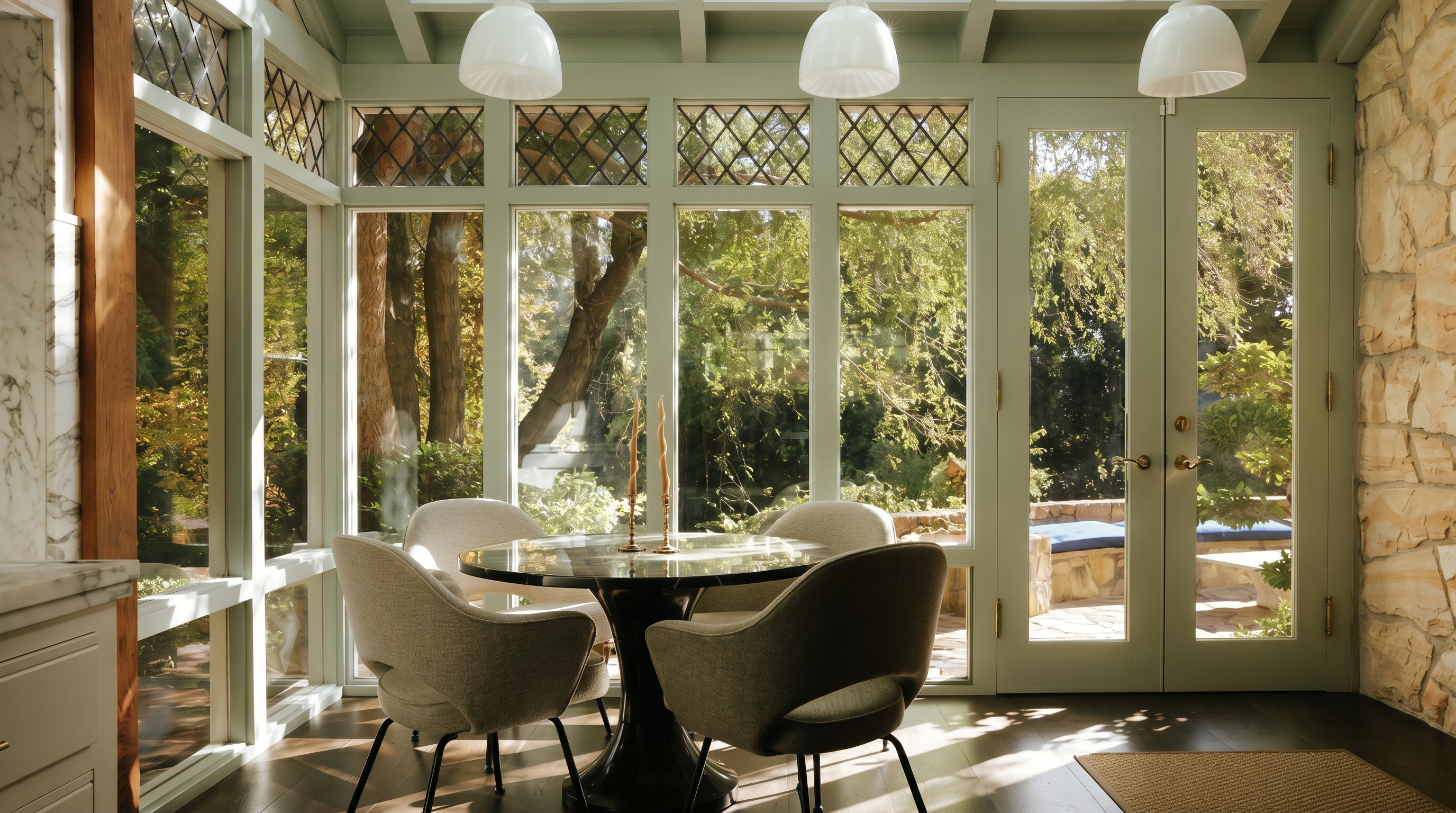 This bright and airy sunroom features a circular dining table surrounded by four modern upholstered chairs, set against a backdrop of floor-to-ceiling windows and glass-paned doors. The space is accented by a vaulted ceiling with exposed beams, three elegant pendant lights, and a natural stone wall, creating a seamless transition between the interior and the lush outdoor greenery. The perspective is a straight-on, eye-level shot that emphasizes the room's open, tranquil atmosphere and connection to nature.