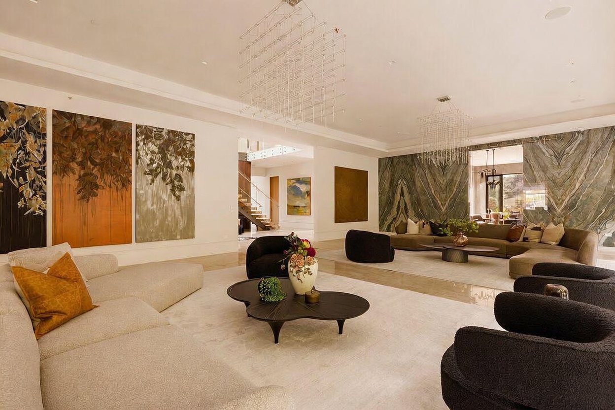 This expansive, open-concept living area features a sophisticated, modern design with a sunken seating section and high ceilings adorned with intricate, geometric light fixtures. The space is anchored by a large, textured stone wall behind a neutral-toned sectional, complemented by a series of large, abstract art pieces on the adjacent wall. The room flows seamlessly into a secondary lounge area, creating a grand and airy atmosphere suitable for high-end entertaining.