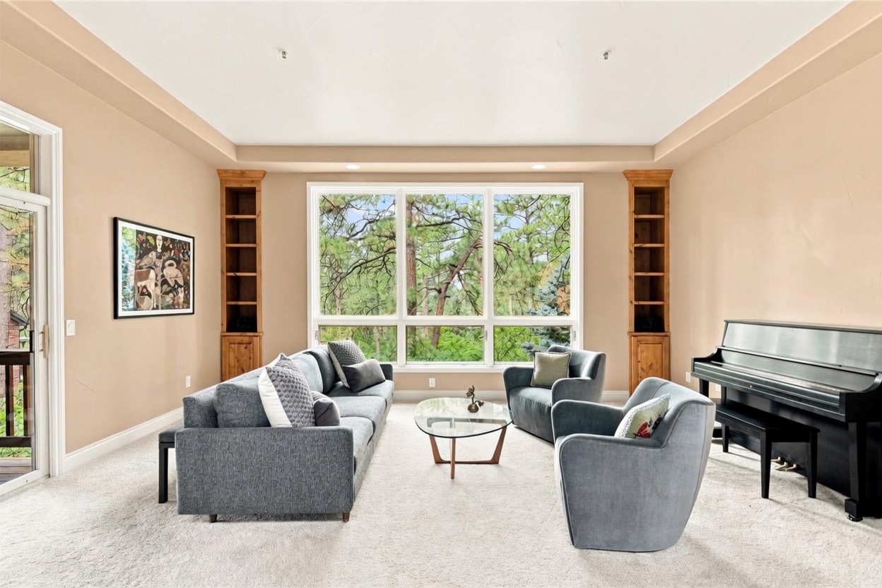 This spacious living room features a large, central window arrangement that provides a clear view of the surrounding trees. The room is furnished with a long grey sofa, two matching armchairs, and a glass-topped coffee table, all arranged on a neutral carpet. To the right, an upright piano sits against the wall, while built-in wooden shelving units flank the large window, creating a balanced and inviting atmosphere.