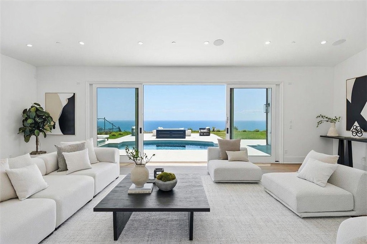 This bright and airy living room features a minimalist, contemporary design with a neutral color palette and plush white sectional seating. A large, dark wood coffee table serves as a central anchor, while expansive sliding glass doors provide a seamless transition to an outdoor pool area with a stunning ocean view. The space is accented by curated artwork and decorative greenery, creating a sophisticated and serene atmosphere.