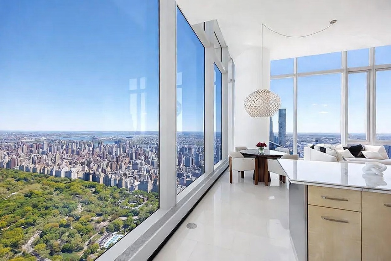 This high-rise interior space features a dining area adjacent to a kitchen, characterized by expansive floor-to-ceiling windows that offer a panoramic view of the city and a large park below. The room is bright and minimalist, with white flooring, a circular dining table, and a distinctive spherical pendant light fixture. The perspective captures the seamless transition between the sleek indoor living area and the dramatic urban landscape outside.