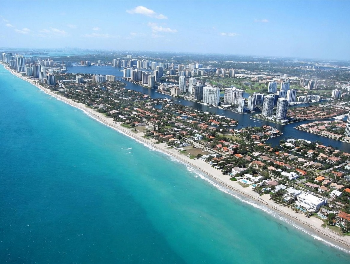 This expansive aerial view captures a stunning coastal landscape featuring a vibrant turquoise ocean meeting a sandy shoreline. The scene showcases a dense residential area with numerous high-rise condominiums and single-family homes nestled along winding waterways and lush green spaces. The perspective provides a comprehensive look at the prime waterfront location, highlighting the proximity of the properties to the beach and the surrounding urban infrastructure.