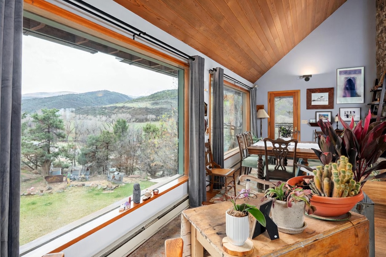 This dining area features a vaulted wood-paneled ceiling and expansive windows that frame a scenic mountain and landscape view. The room is furnished with a wooden dining table and chairs, complemented by a rustic wooden console table in the foreground adorned with potted plants and a cactus. The space offers a warm, inviting atmosphere with a seamless connection to the natural surroundings outside.