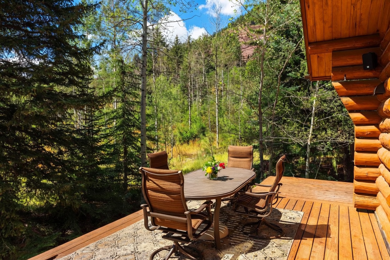 This elevated wooden deck offers a serene outdoor dining space surrounded by a lush forest of aspen and pine trees. The area is furnished with a metal-framed dining set and features a decorative metal railing with a branch-like design. The perspective captures the inviting atmosphere of the deck, highlighting the natural beauty of the mountain landscape beyond.