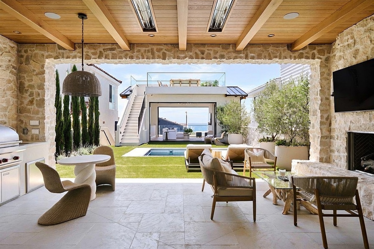 This luxurious covered outdoor living space features a stone-clad chimney breast, a built-in grill station, and a dining area, all sheltered by a wood-beamed ceiling. The space opens directly onto a manicured lawn with a swimming pool, leading the eye toward a secondary structure with a rooftop deck and ocean views. The design seamlessly blends indoor comfort with an open-air coastal aesthetic, creating an ideal environment for high-end entertaining.