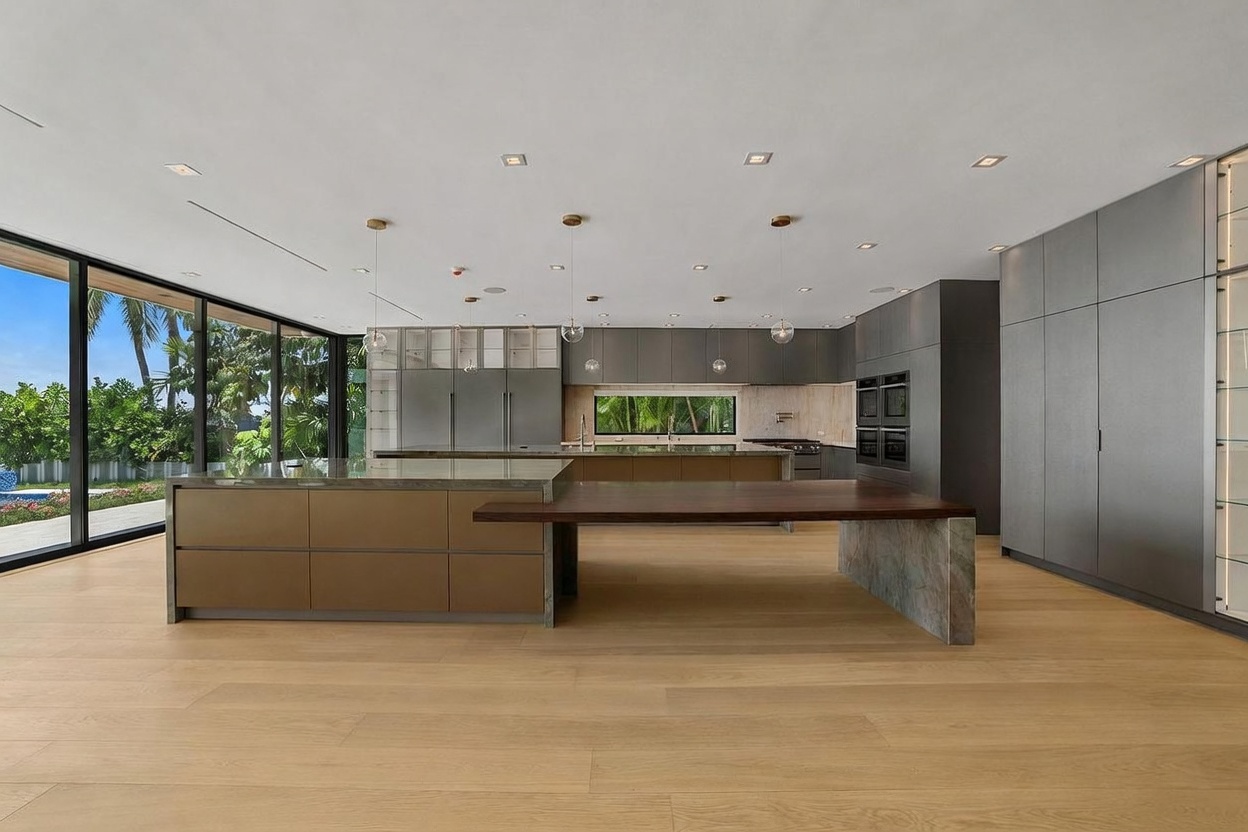 This expansive, modern kitchen features a large central island with a dark wood dining extension and a secondary island behind it, both topped with stone surfaces. The space is defined by sleek, dark grey cabinetry, integrated appliances, and light wood flooring that creates a warm contrast. Large, floor-to-ceiling windows along the left wall provide an abundance of natural light and a seamless view of the lush outdoor greenery.