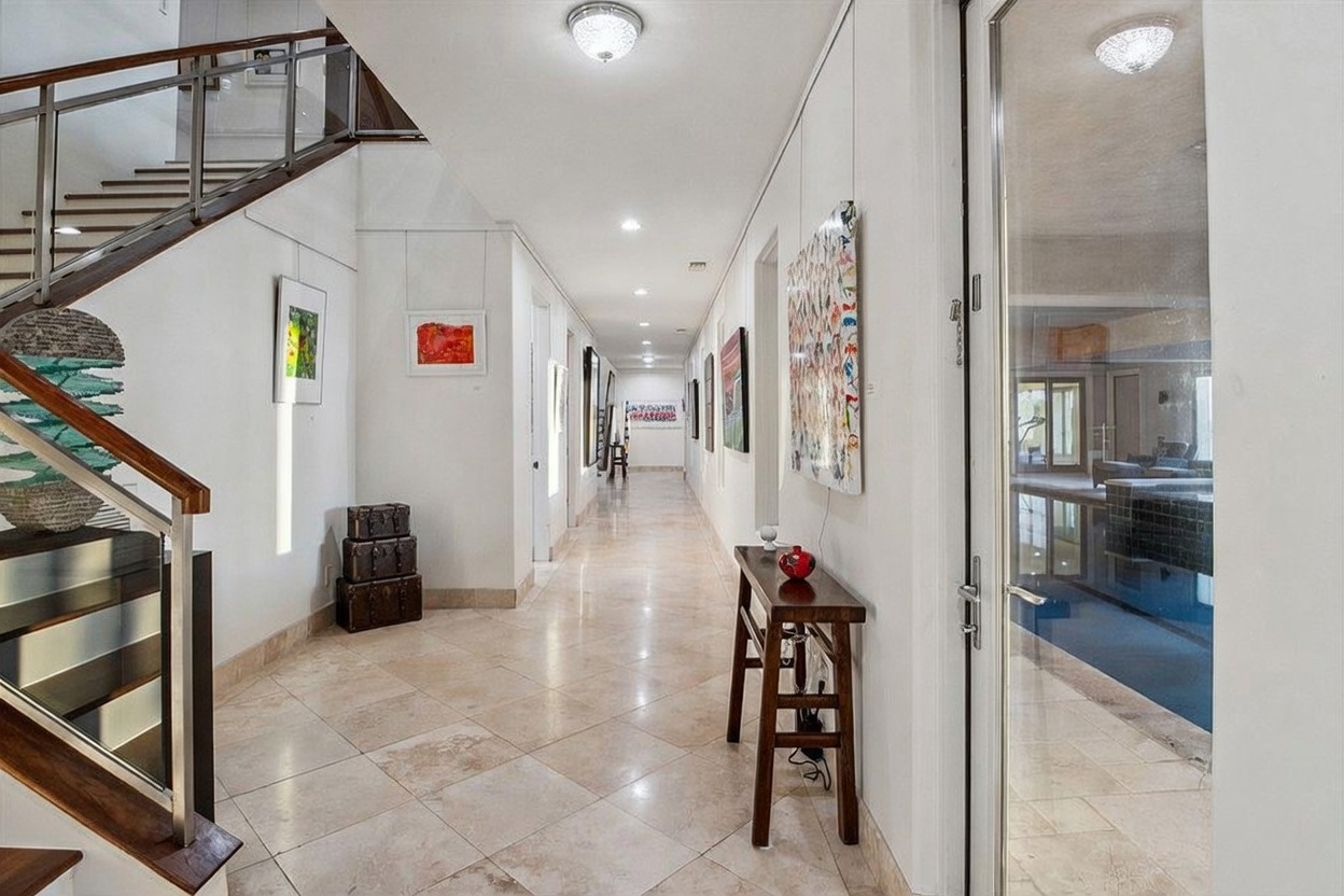 This spacious, light-filled hallway features polished stone flooring and a grand staircase with a modern metal and wood railing on the left. The long corridor is lined with white walls adorned with various framed artworks, leading the eye toward the back of the home. To the right, a glass-paneled door offers a view into an indoor pool area, creating a bright and airy atmosphere throughout the space.