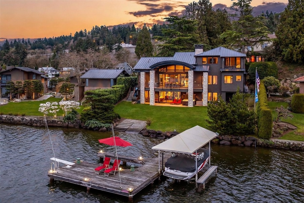 This stunning waterfront property features a multi-level contemporary home with dark siding and prominent stone accents. The rear of the house showcases large windows and glass doors that open onto a spacious lawn, leading directly to a private wooden dock equipped with a boat lift and a red umbrella. The scene is captured at sunset, casting a warm, golden glow over the tranquil water and the meticulously landscaped grounds.