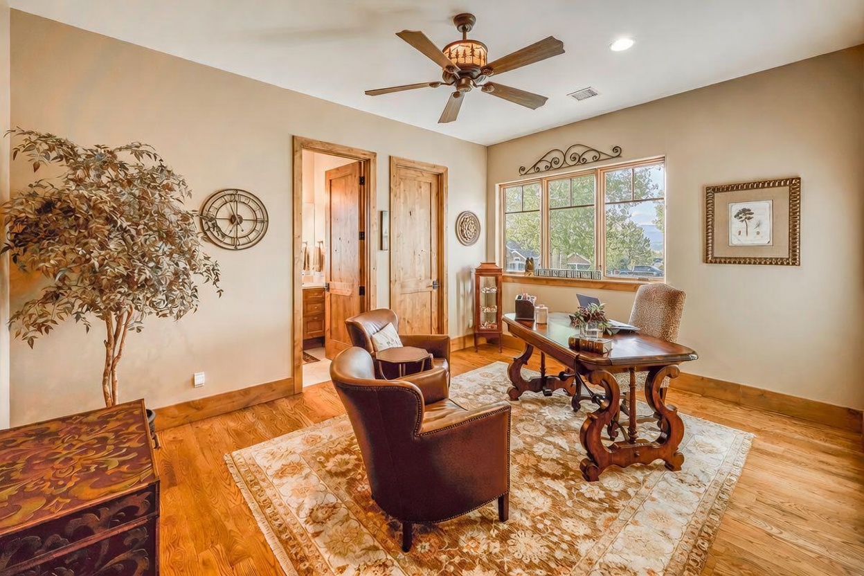 This elegant home office features warm-toned wood flooring, a large central desk, and two leather armchairs arranged on a patterned area rug. The room is accented by a decorative wall clock, a potted tree, and large windows that provide natural light and a view of the outdoors. The space is finished with wood trim and a ceiling fan, creating a sophisticated and comfortable work environment.
