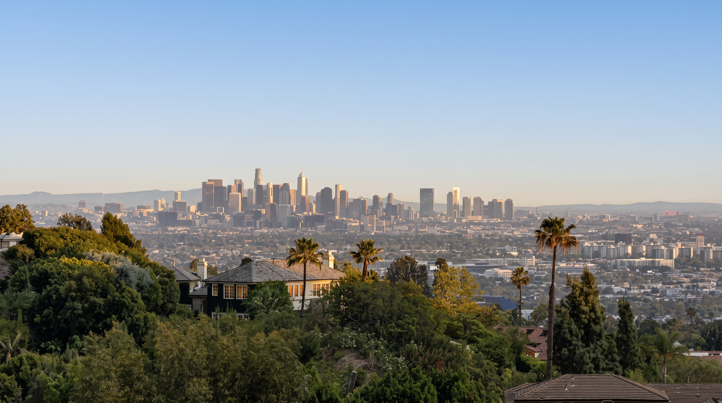 This elevated perspective captures a sweeping, panoramic view of the Los Angeles skyline set against a clear blue sky. In the foreground, lush green trees and the tiled roofs of residential homes frame the vista, emphasizing the property's prime hillside location. The composition highlights the contrast between the dense urban cityscape in the distance and the serene, verdant landscape of the immediate neighborhood.
