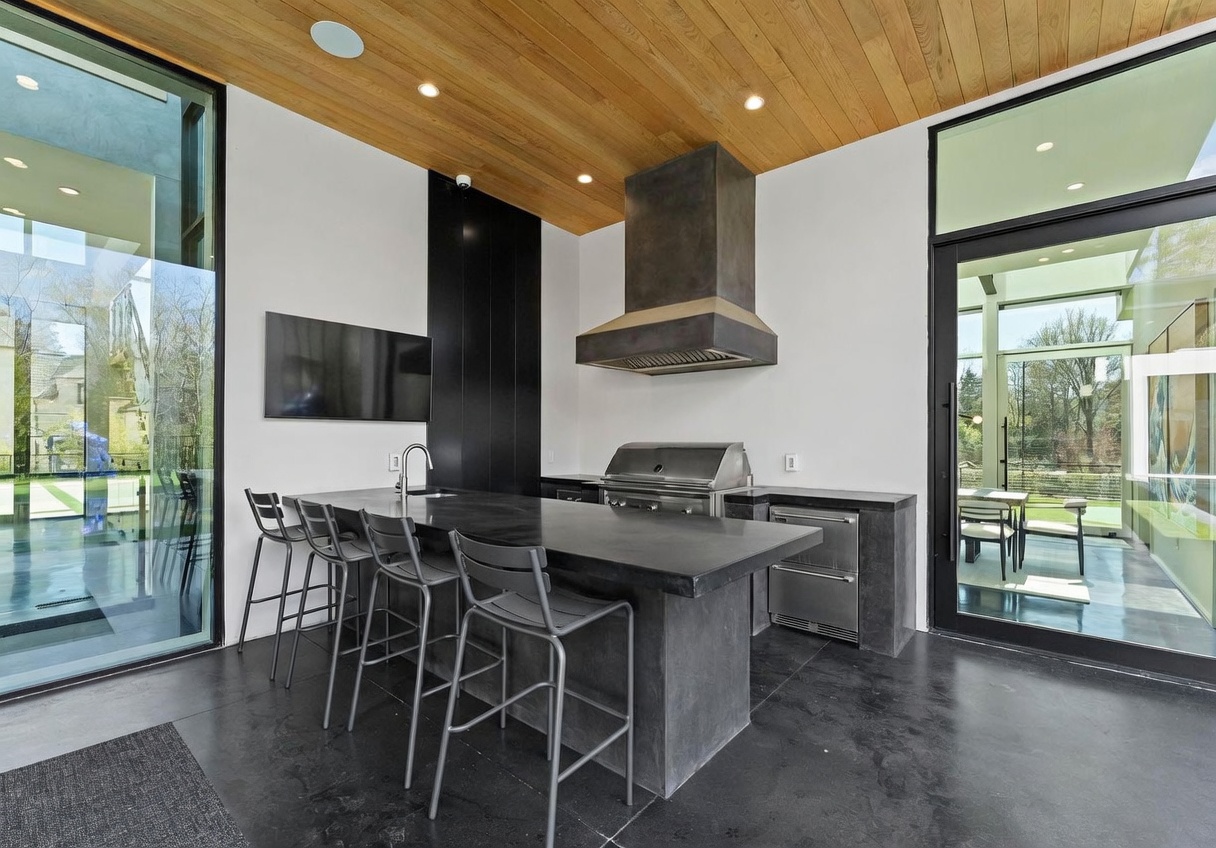This modern outdoor kitchen area features a sleek, dark stone island with integrated seating and a stainless steel grill setup. The space is defined by a warm wood-paneled ceiling and large glass walls that provide a seamless transition to the outdoor environment. The minimalist design, combined with the dark color palette and clean lines, creates a sophisticated and contemporary atmosphere for outdoor entertaining.