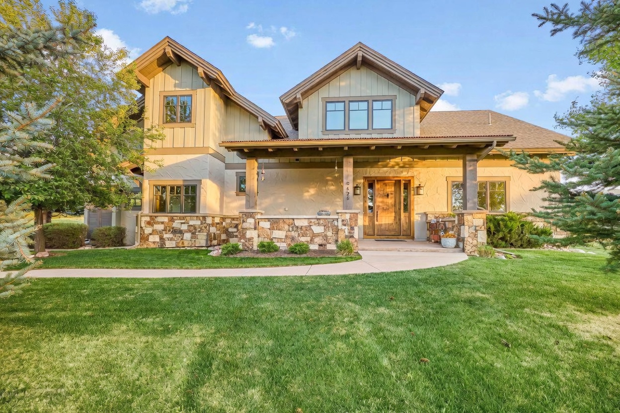 This front-facing view captures a custom mountain-style home featuring a blend of light-toned stucco and vertical wood siding. The residence is highlighted by a covered front porch with stone-clad pillars and a prominent wooden front door, set against a backdrop of mature evergreen trees and a manicured lawn. The architectural design includes multiple gables and a warm, inviting aesthetic that emphasizes natural textures and curb appeal.