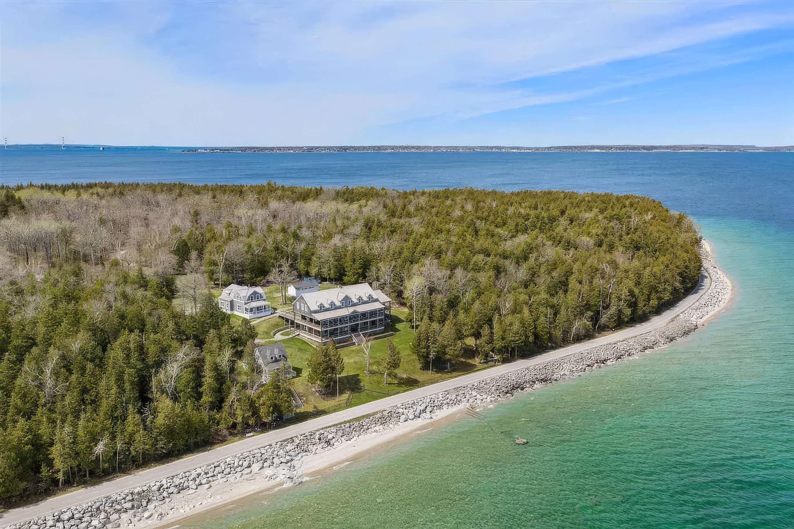 This high-angle aerial view captures a secluded waterfront property nestled within a dense forest on a peninsula. The main residence features a multi-level design with expansive wrap-around porches, accompanied by a smaller secondary structure and a detached garage. The property is bordered by a rocky shoreline and clear turquoise water, offering a serene and private coastal setting.