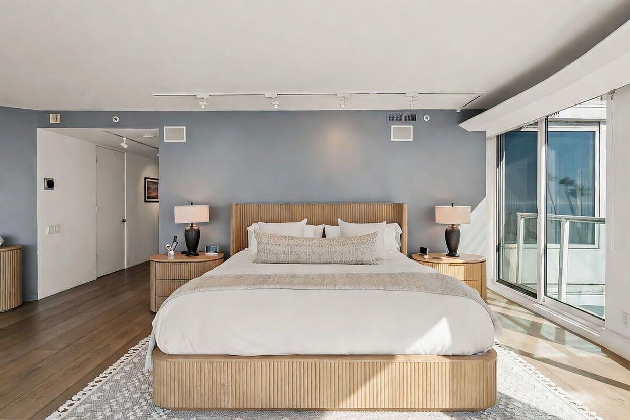 This primary bedroom features a contemporary design with a light wood bed frame and matching nightstands set against a soft blue accent wall. Large sliding glass doors provide ample natural light and a view of the outdoors, while a patterned area rug adds texture to the hardwood flooring. The room is illuminated by track lighting and bedside lamps, creating a serene and sophisticated atmosphere.