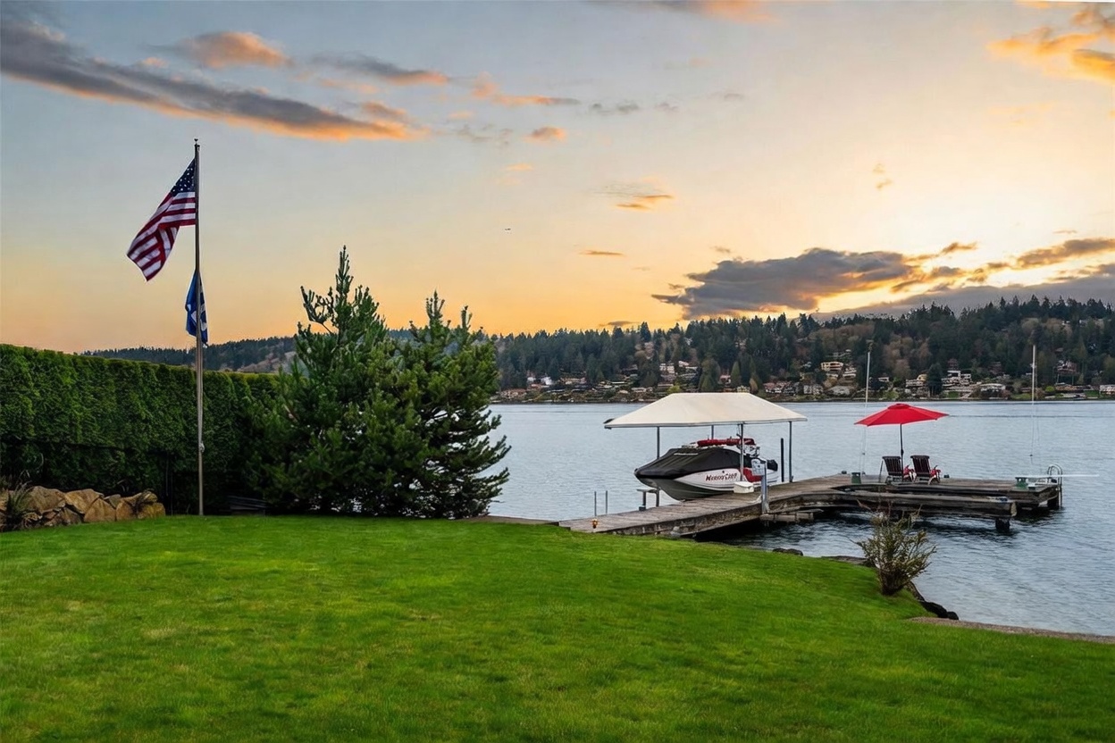 This serene waterfront property features a lush, manicured green lawn that leads directly to a private wooden dock. The scene includes a boat under a canopy, a red patio umbrella with chairs, and an American flag waving in the breeze, all set against a picturesque sunset over the water and distant treeline. The perspective is a wide, eye-level shot capturing the tranquil transition from the private yard to the expansive lakefront.