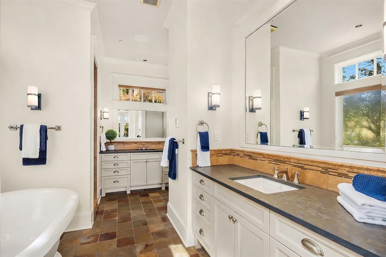 This spacious primary bathroom features a dual-vanity layout with dark stone countertops, white cabinetry, and a large wall-to-wall mirror. A deep soaking tub sits to the left, while the room is finished with warm-toned slate tile flooring and a decorative tile backsplash. The perspective captures the depth of the room, highlighting the bright, clean aesthetic and the transition between the two vanity areas.