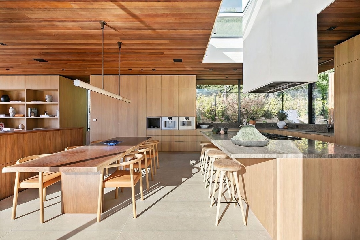 This sophisticated, modern kitchen features a seamless blend of natural wood cabinetry and a large stone-topped island, creating a warm and inviting atmosphere. A striking wooden ceiling with integrated lighting and a skylight above the range hood adds architectural depth, while the open-concept layout flows effortlessly into a dining area with a custom live-edge table. The space is bathed in natural light, highlighting the high-end finishes and the serene view of the lush greenery outside.
