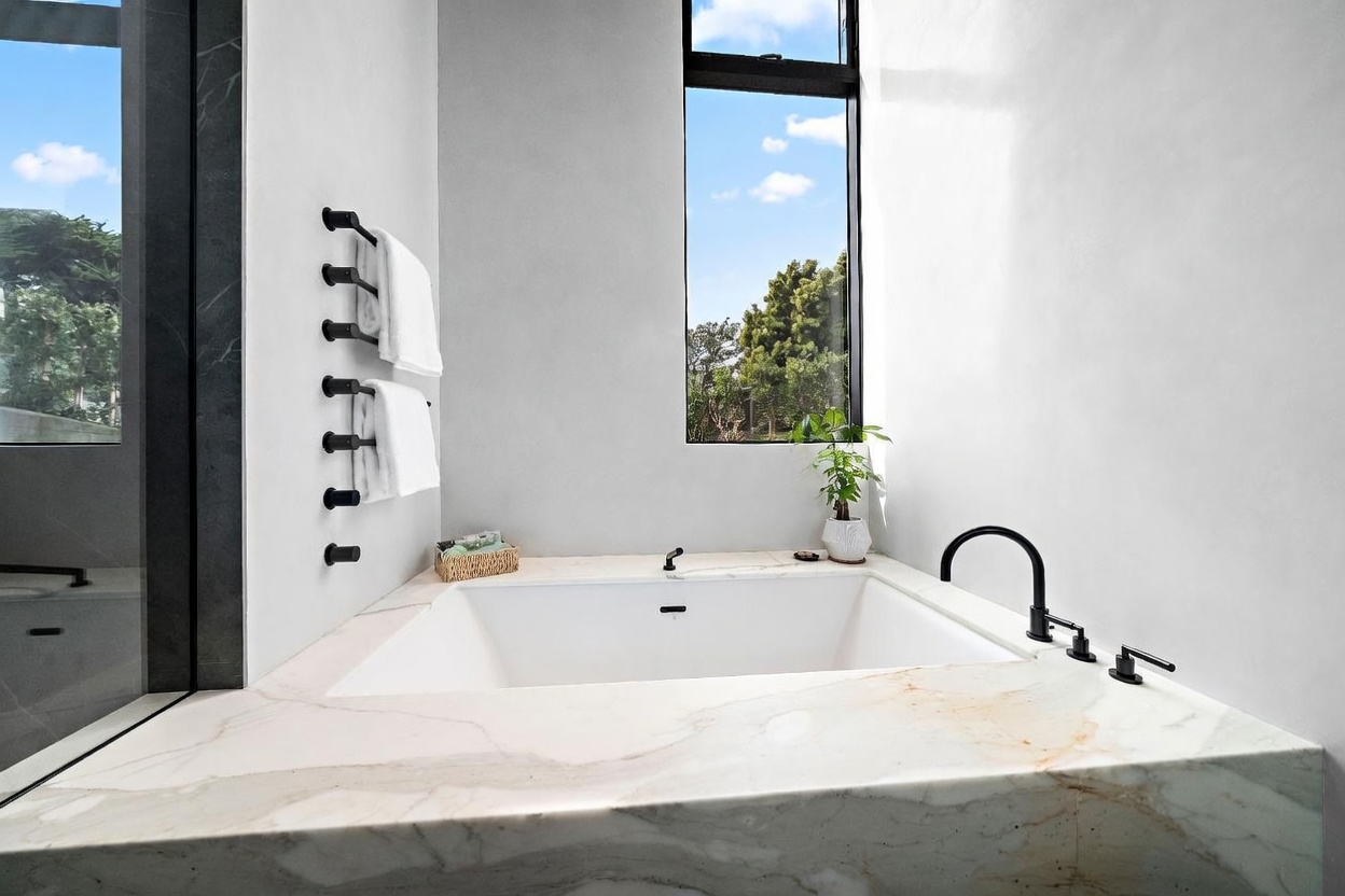 This luxurious primary bathroom features a deep soaking tub encased in a light-colored stone surround with prominent veining. A tall, narrow window provides natural light and a view of the outdoors, while matte black fixtures add a modern contrast. The space is minimalist and serene, with white towels hanging on a wall-mounted rack and a small potted plant adding a touch of greenery.