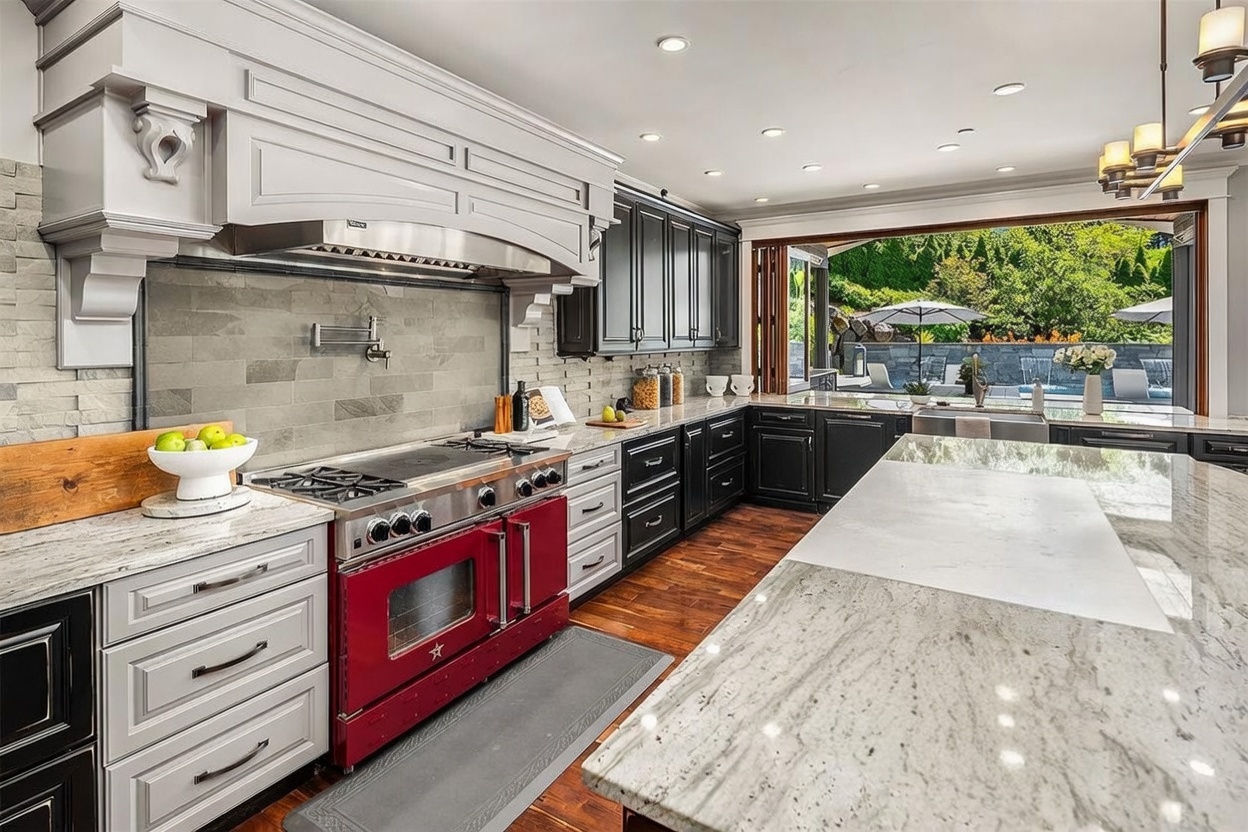 This high-end kitchen features a striking red range as a focal point, complemented by a mix of light and dark cabinetry and stone countertops. A large island with a stainless steel sink sits in the foreground, while a wide opening with folding doors provides a seamless transition to an outdoor patio and pool area. The space is illuminated by recessed lighting and offers a sophisticated, functional design with a view of the lush backyard.