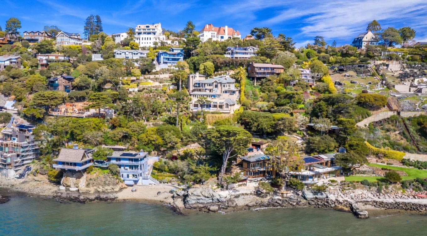 This aerial perspective captures a dramatic coastal hillside community featuring a collection of multi-level luxury homes nestled into a steep, tree-covered slope overlooking the water. The architecture varies from modern glass-fronted residences to traditional hillside estates, all connected by winding paths and lush, terraced landscaping. The scene emphasizes the prime waterfront location, showcasing the rugged shoreline and the expansive, elevated views enjoyed by the properties.