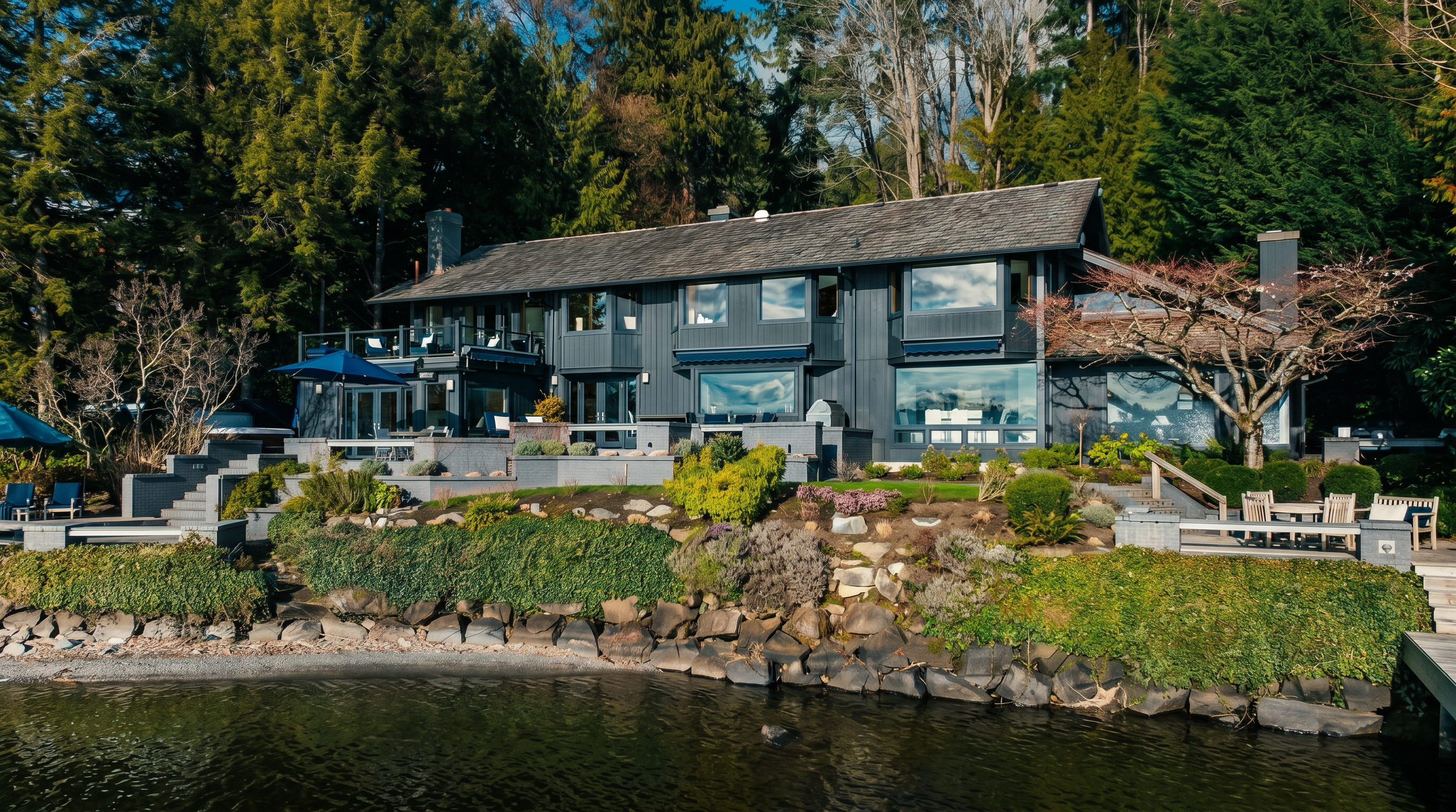This stunning waterfront property features a multi-level dark-toned home nestled against a lush, forested backdrop. The rear exterior showcases expansive tiered stone patios, a hot tub, and large windows that offer panoramic views of the lake, creating a serene and luxurious atmosphere. The landscaping is meticulously terraced with stone retaining walls, leading down to the natural rocky shoreline.