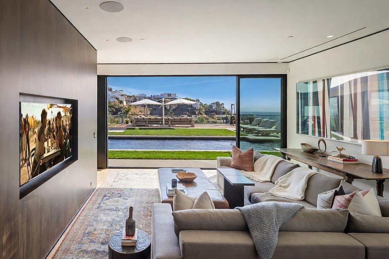 This contemporary living room features a large, comfortable sectional sofa and a low-profile coffee table, all oriented toward a wall-mounted television. The space seamlessly transitions to an outdoor area through expansive glass sliding doors that reveal a swimming pool, a manicured lawn, and a scenic coastal view. The interior design is characterized by a neutral color palette, a patterned area rug, and a long console table adorned with art, creating a sophisticated and airy atmosphere.