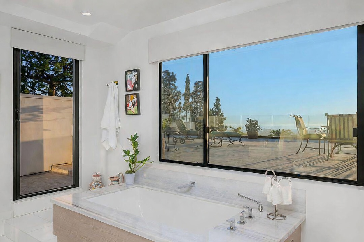 This bright primary bathroom features a deep soaking tub set within a light-toned stone surround, positioned directly beneath a large window that offers expansive views of an outdoor deck. The room is bathed in natural light, highlighting the clean, minimalist aesthetic and the seamless transition between the indoor space and the exterior terrace. Two framed pieces of art hang on the wall, adding a touch of personality to the otherwise serene and neutral environment.