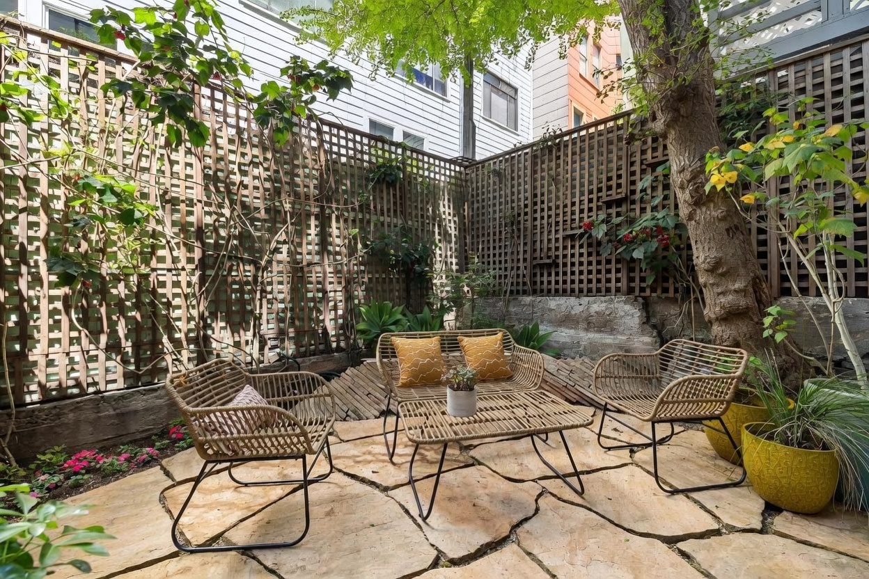 This charming, secluded garden patio features natural stone pavers arranged in a flagstone pattern, creating a rustic and inviting outdoor seating area. The space is furnished with a matching set of woven wicker-style chairs and a low coffee table, accented by vibrant yellow throw pillows. Enclosed by tall wooden lattice fencing and lush greenery, the area offers a private, tranquil retreat shaded by a mature tree.