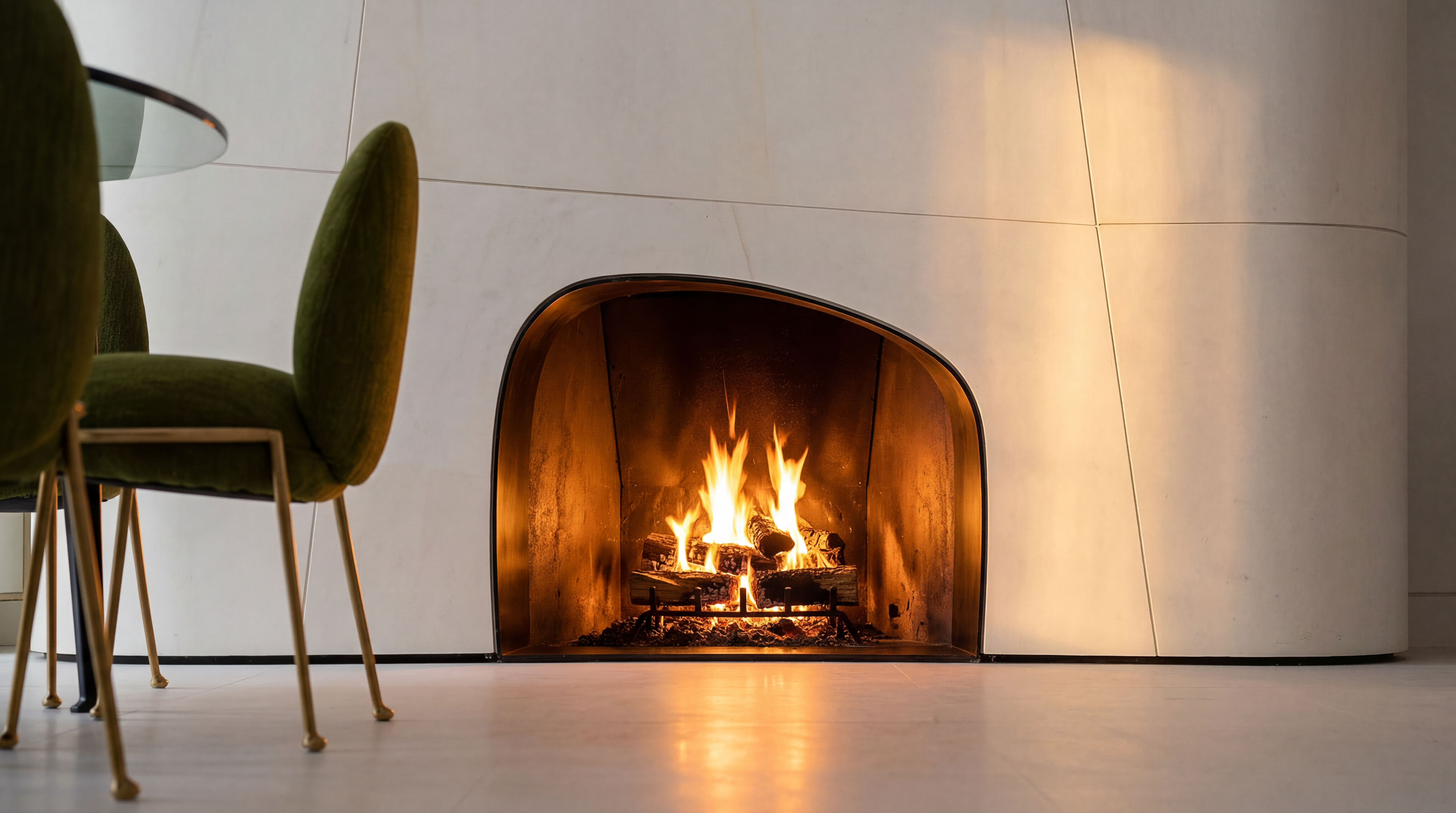 This interior shot highlights a stunning, modern fireplace with a unique, organic-shaped hearth set into a seamless, oversized light-colored wall panel. A green upholstered chair with elegant thin legs sits to the left, adding a pop of texture and color. The warm glow from the flames against the sculptural wall creates an inviting and sophisticated atmosphere, characteristic of a high-end luxury home design.