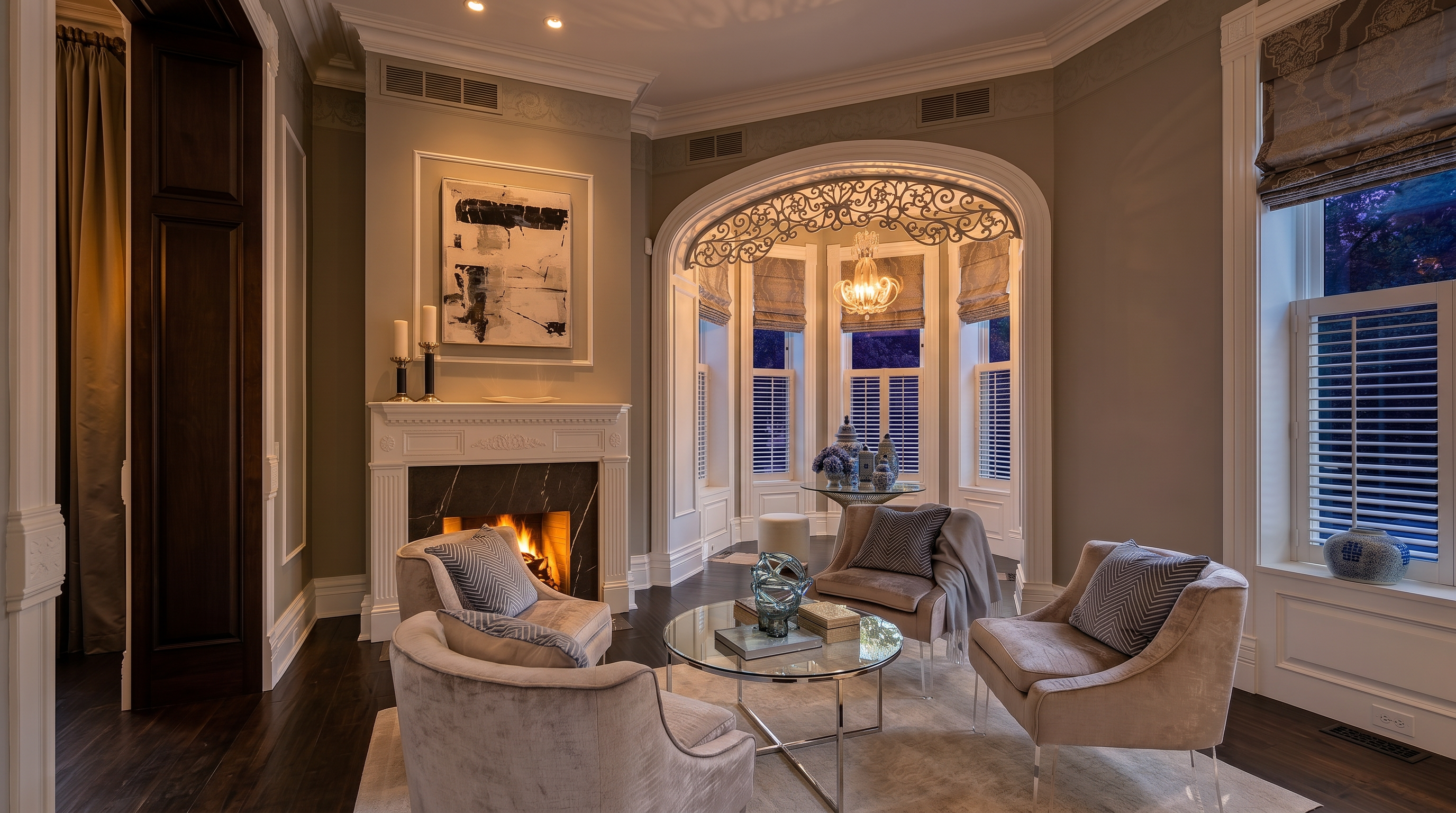 This elegant living room features a sophisticated neutral color palette with dark wood flooring and a prominent fireplace with a dark stone surround. The space is furnished with four plush armchairs arranged around a circular glass coffee table, creating an inviting conversation area. A large, arched window bay with intricate decorative metalwork above the windows allows for abundant natural light, enhancing the refined and airy atmosphere of the room.