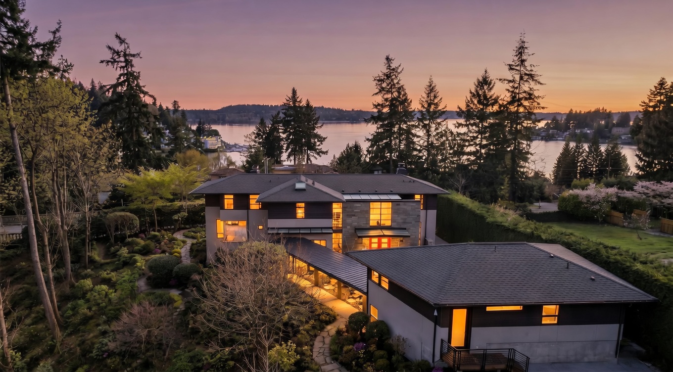 This aerial perspective captures a contemporary multi-level residence at dusk, set against a backdrop of a serene lake and a vibrant purple and orange sunset. The home features a mix of stone and dark siding, connected by a covered walkway that leads to a separate garage structure, all surrounded by lush, landscaped gardens and mature trees. The warm glow from the interior lighting contrasts beautifully with the cool tones of the twilight sky, highlighting the property's sophisticated architectural design.