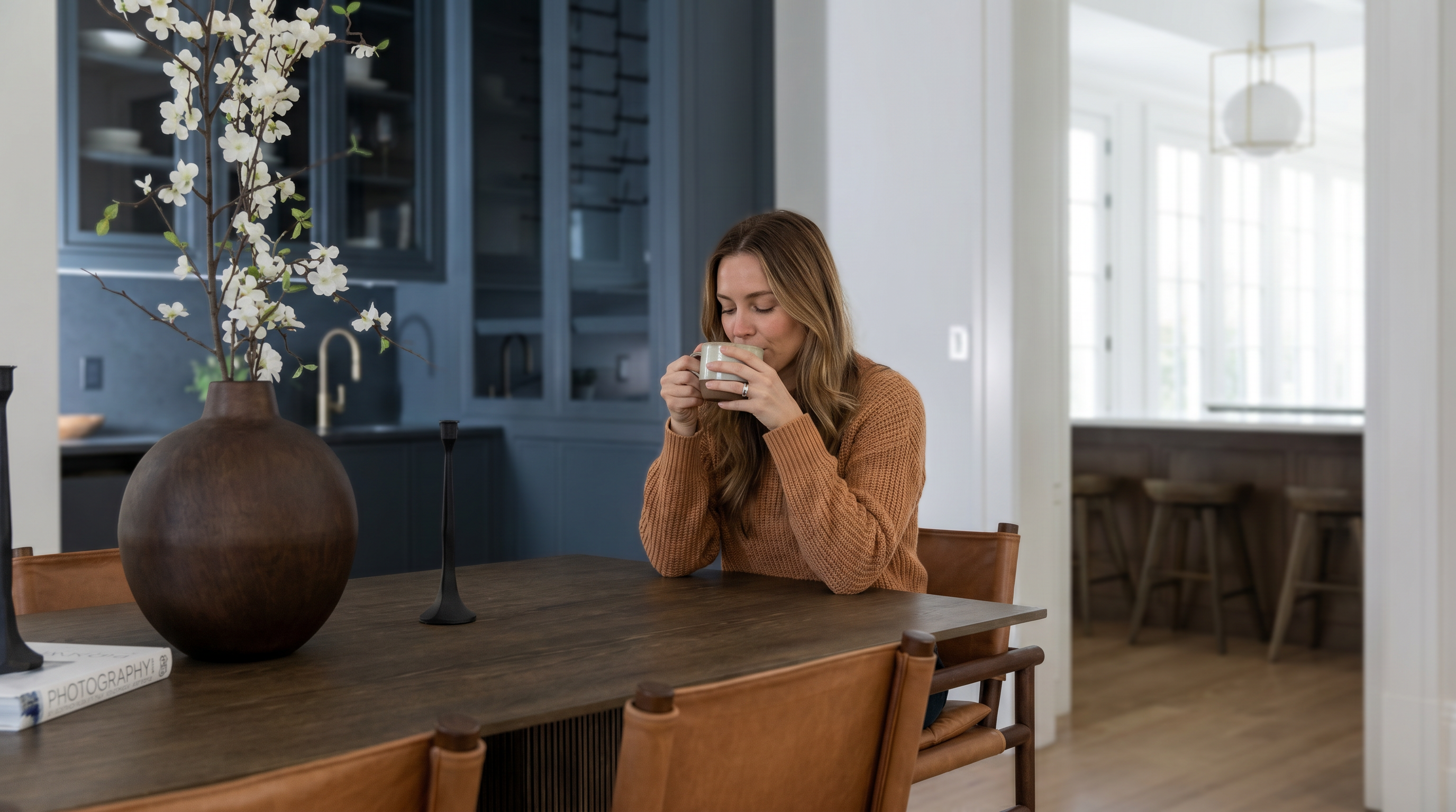 This elegant dining area features a dark wood table surrounded by leather-upholstered chairs, creating a sophisticated and warm atmosphere. In the background, a built-in dark blue cabinetry unit serves as a stylish bar or storage area, while an open doorway provides a glimpse into a kitchen with bar seating. The composition uses a shallow depth of field to focus on the dining table's centerpiece, emphasizing the refined textures and modern aesthetic of the home.